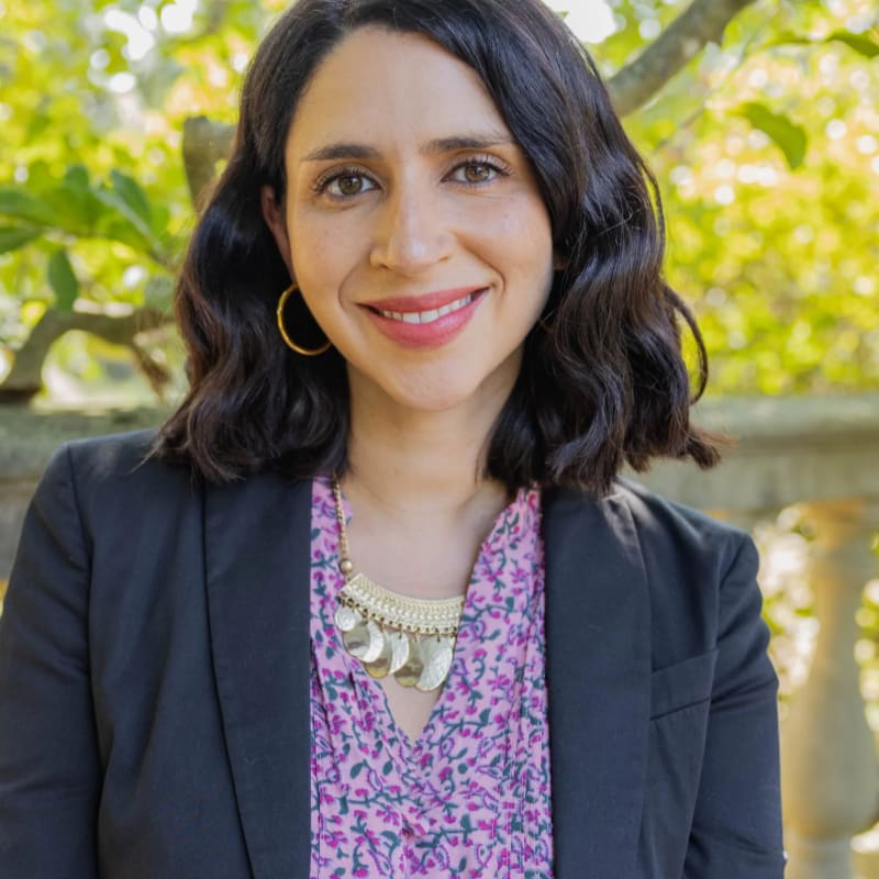 A woman with dark hair and a warm smile is in the foreground, wearing a black blazer over a pink floral shirt and a gold necklace, while a blurred background of green foliage and a stone balustrade suggests an outdoor setting.