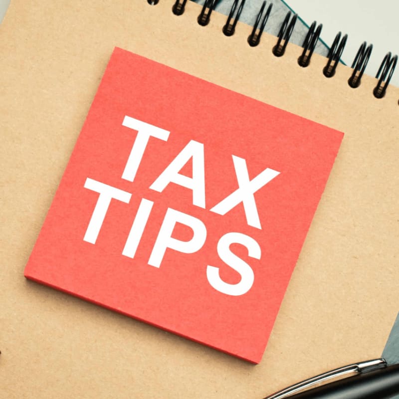 A bright red sticky note with "TAX TIPS" in white letters rests on a brown notebook, with a pen visible in the lower right corner.