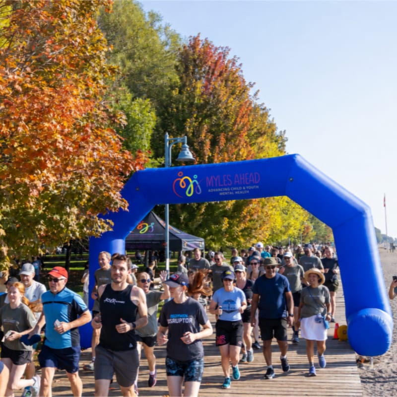 A group of people run through a blue inflatable archway on a wooden boardwalk, with colorful autumn trees and a beach in the background.