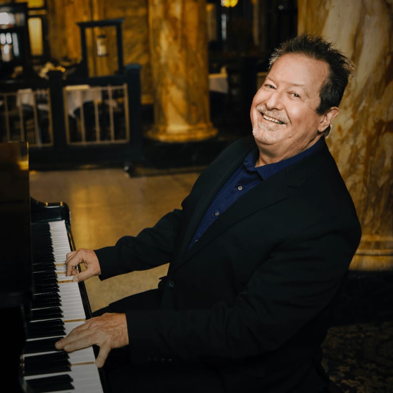 A smiling man in a suit plays a piano in the foreground, with a dimly lit, ornate room in the background.