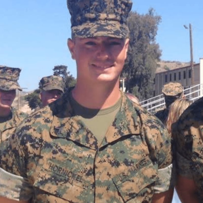A young man in a Marine uniform smiles in the foreground, with other Marines and buildings in the background under a clear sky.