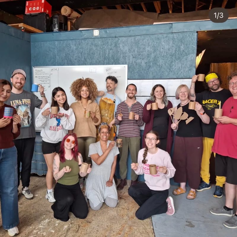 A group of people stand in a workshop, holding cups, with a whiteboard and tools visible in the background.