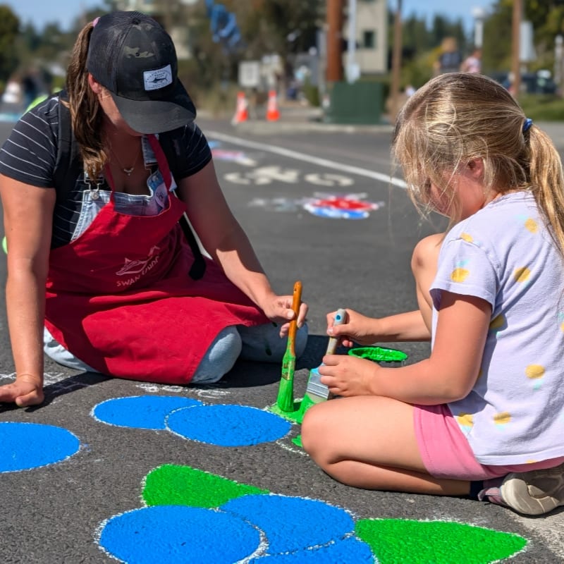 A woman in a red apron and a young girl with blonde hair paint blue and green circles on a street, with blurred buildings and trees in the background.