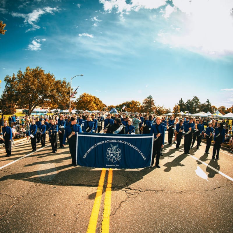 A marching band in blue uniforms parades down a street carrying a banner, with spectators and trees lining the background under a bright sky.