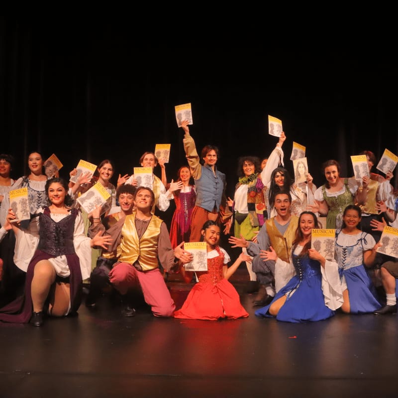 A group of actors in costume on a stage hold up books, with a dark curtain in the background.
