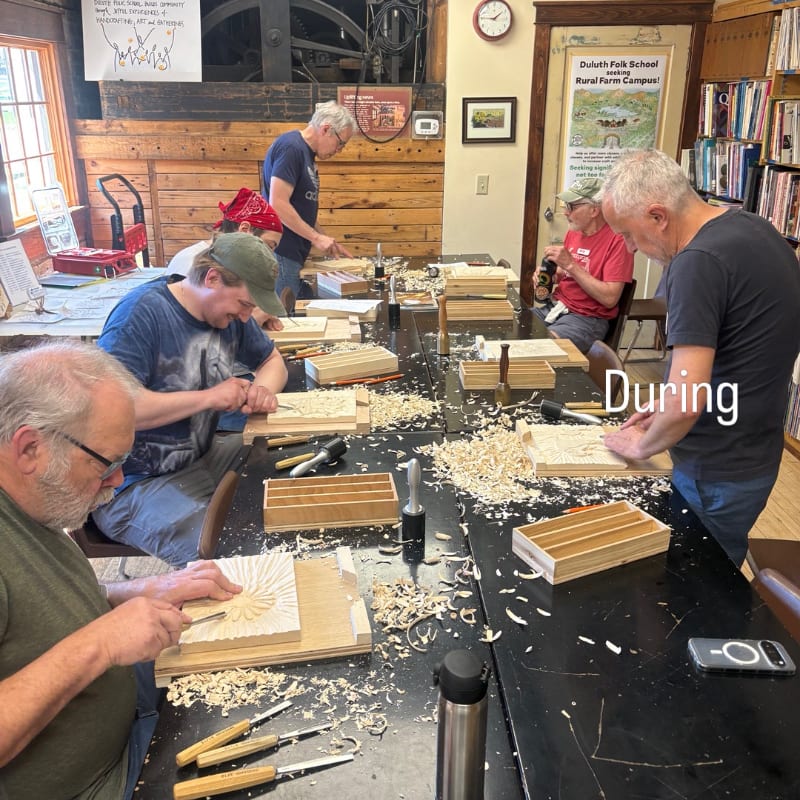 Several men are actively carving wood at a table covered in wood shavings, with shelves of books and a framed poster visible in the background.