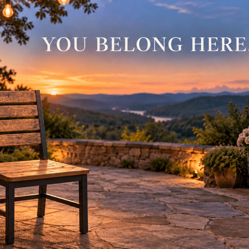A wooden chair sits on a stone patio in the foreground, overlooking a serene landscape of rolling hills and a lake at sunset in the background.