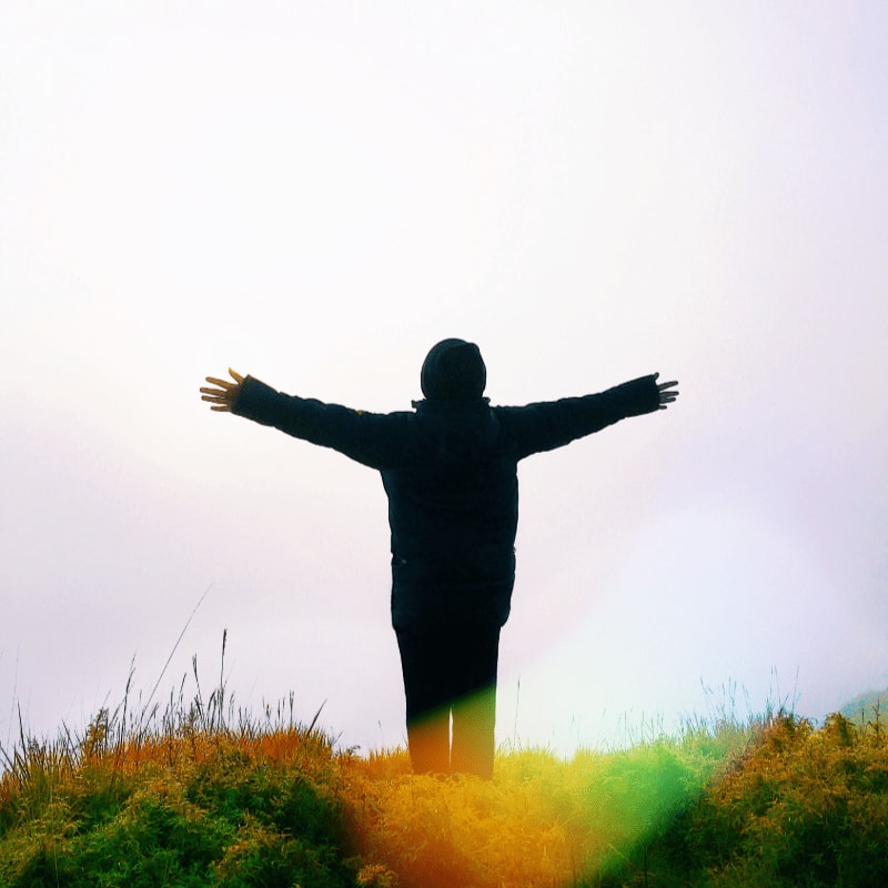 A person in a dark jacket stands with arms outstretched on a grassy hill, silhouetted against a bright, hazy sky.