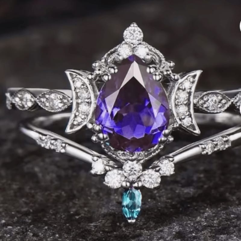 A pear-shaped purple gemstone ring with diamond accents sits on a dark, textured surface, with a matching band featuring crescent moon designs.