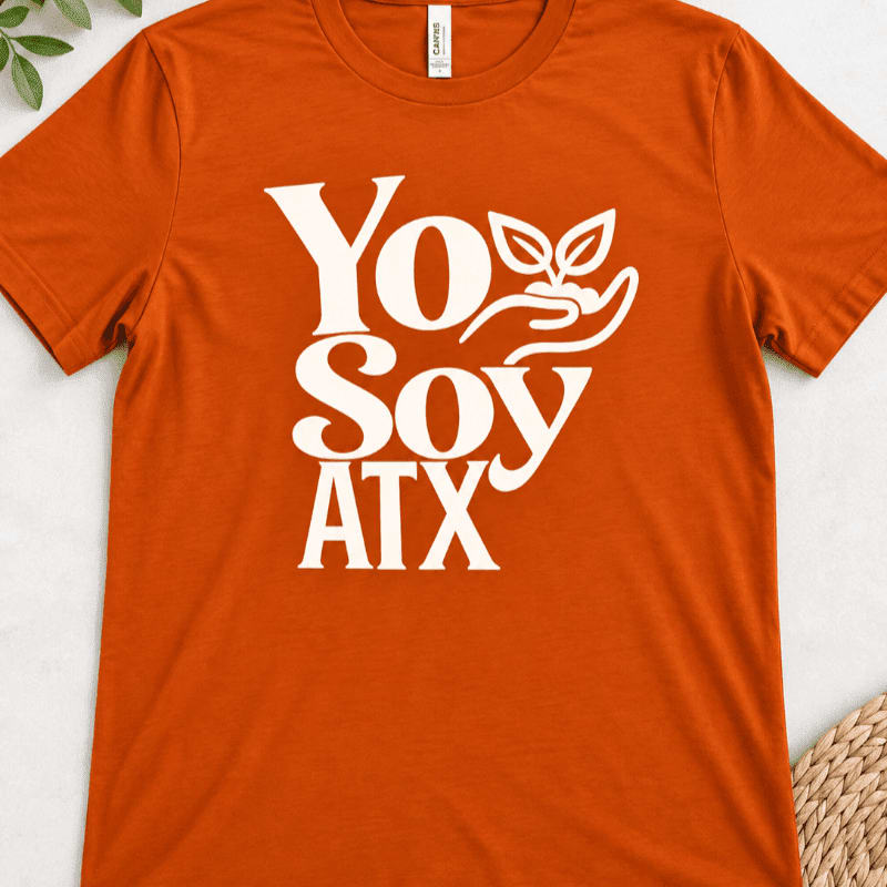 An orange t-shirt with the text "Yo Soy ATX" and a graphic of a hand holding a sprouting plant is laid flat on a light-colored surface with a woven basket in the lower right corner.