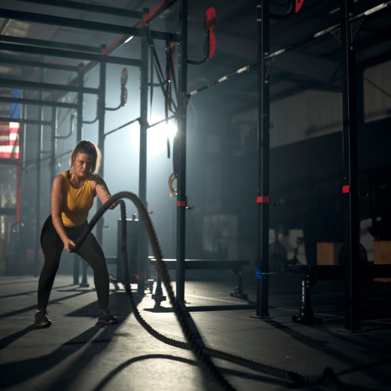 A woman in the foreground is intensely working out with battle ropes in a dimly lit gym, while the background features gym equipment and a hazy American flag.
