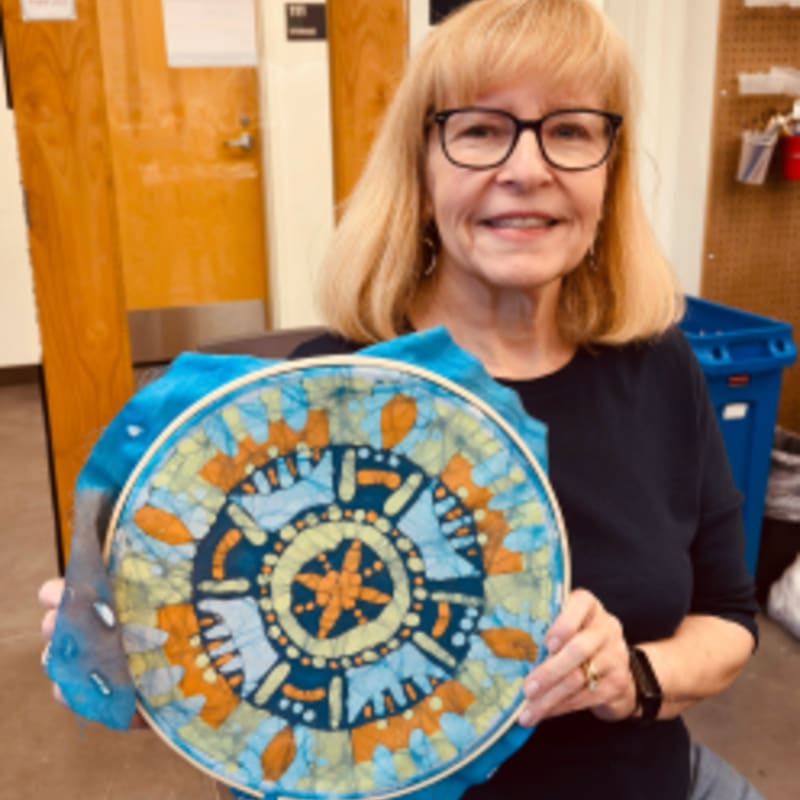 A smiling woman in the foreground holds a colorful batik fabric art piece, while the background shows a classroom or workshop setting with shelves and a blue bin.