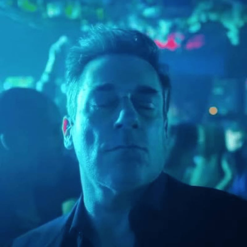 A man with his eyes closed is illuminated by blue light in the foreground, while a blurred crowd dances in the background.