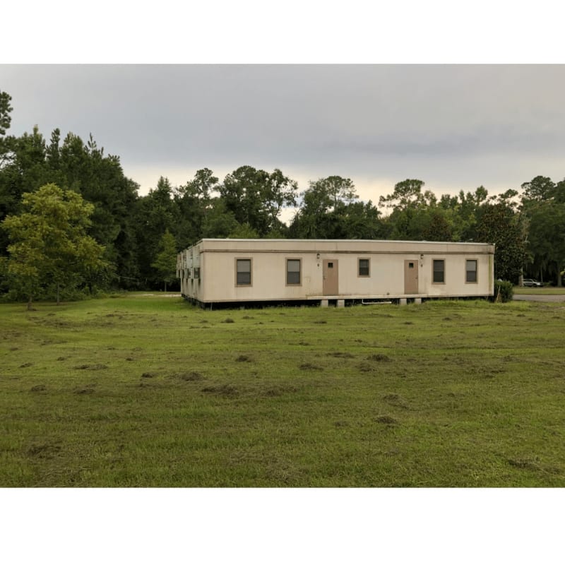 A long, white modular building sits in a mowed field with scattered clumps of grass, backed by a dense line of trees under a cloudy sky.
