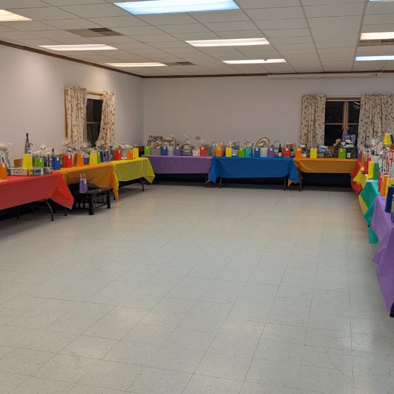 In the foreground, a large, empty tiled floor leads to a room filled with tables draped in colorful cloths, laden with gifts and decorations, while the background shows windows with curtains and fluorescent lighting.