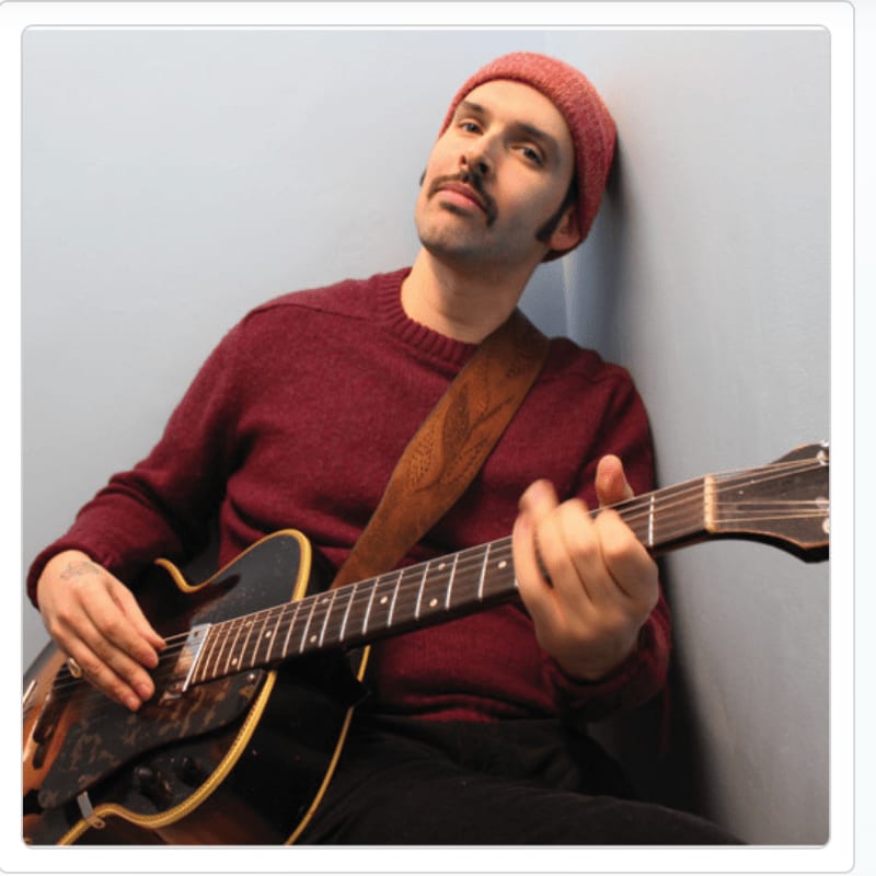 A man in a red beanie and sweater plays an acoustic guitar in front of a light blue wall.