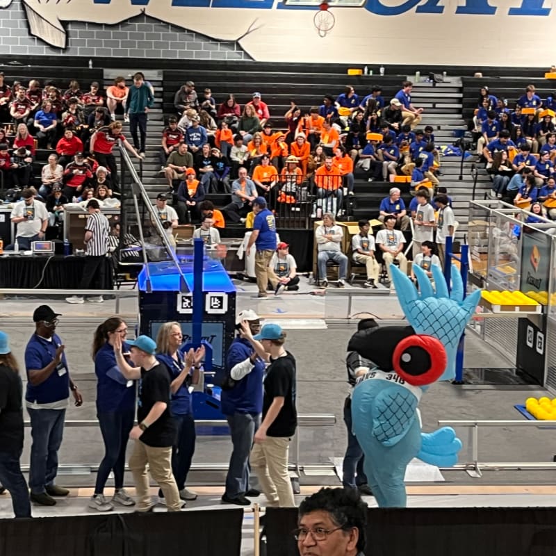 In the foreground, a group of people in blue shirts and hats are gathered around a blue robot, while in the background, a large crowd watches from bleachers in a gymnasium.
