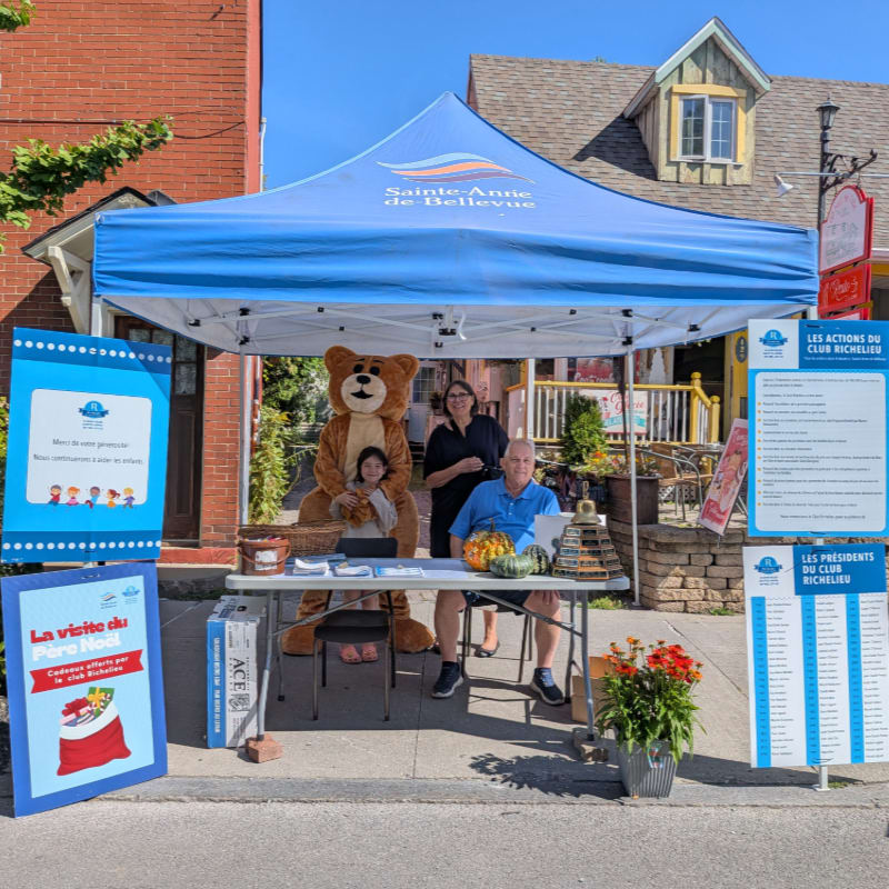 A teddy bear and two people stand behind a table under a blue tent in the foreground, while a street with buildings is visible in the background.