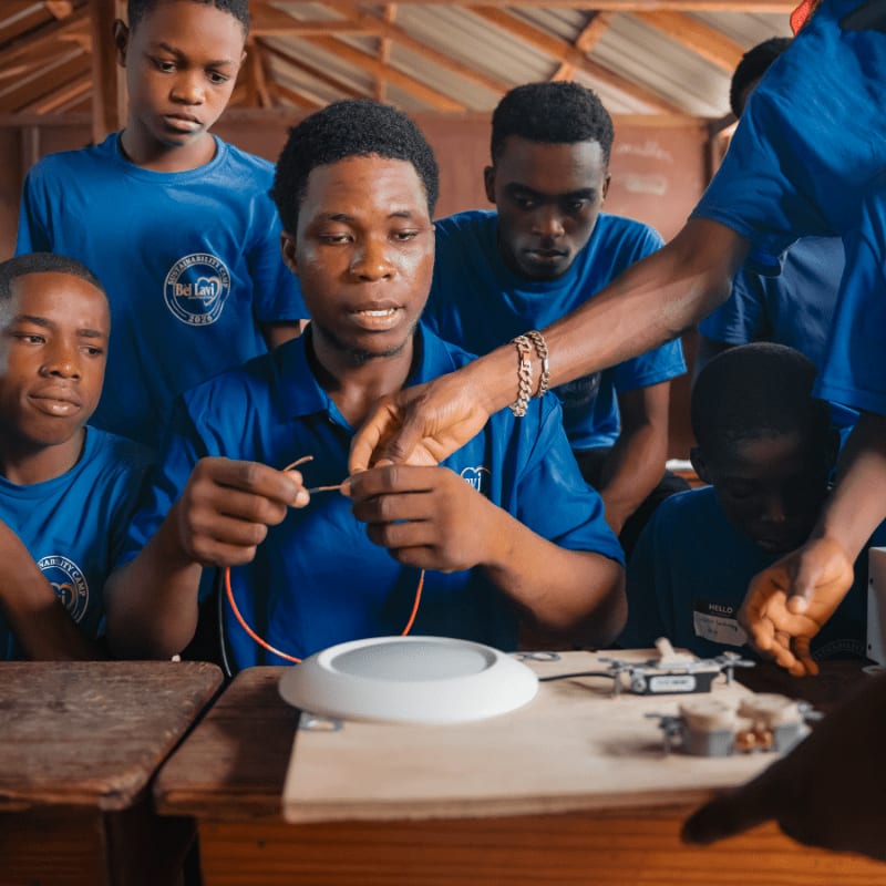 In the foreground, a young man in a blue shirt demonstrates electrical wiring to a group of attentive boys, while the background shows a rustic classroom setting.