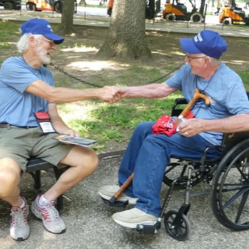 Two men in blue caps shake hands, one seated in a wheelchair with a cane, while forklifts are visible in the blurred background.