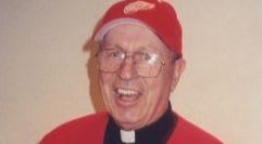 A smiling man in a red baseball cap and clerical collar is pictured against a plain background.