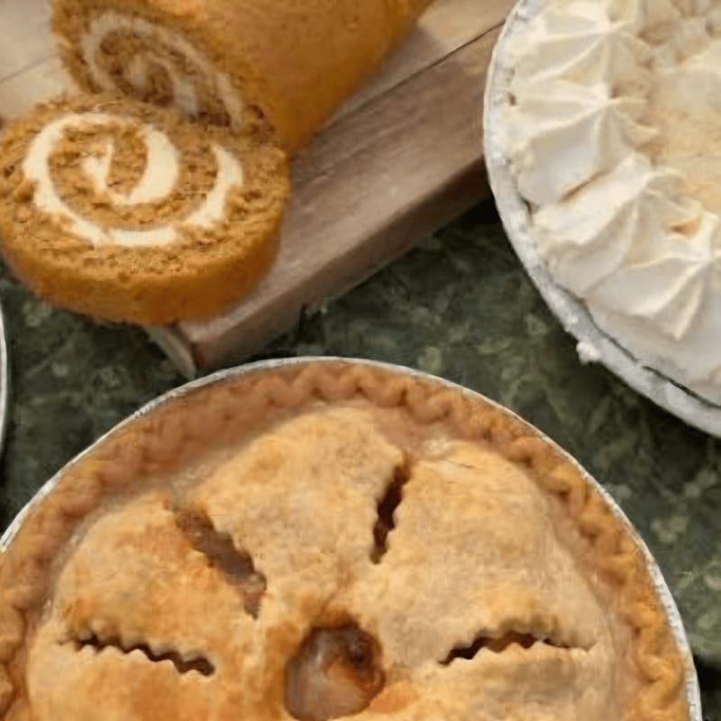A golden-crusted apple pie and a creamy white pie sit on a green patterned surface, with slices of a pumpkin roll on a wooden board in the background.