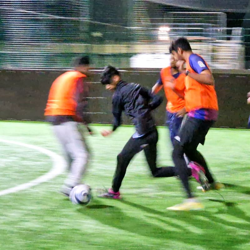 In the foreground, blurred figures in orange vests and dark athletic wear play soccer on a green turf field, while the background shows a dimly lit indoor facility with netting and lights.