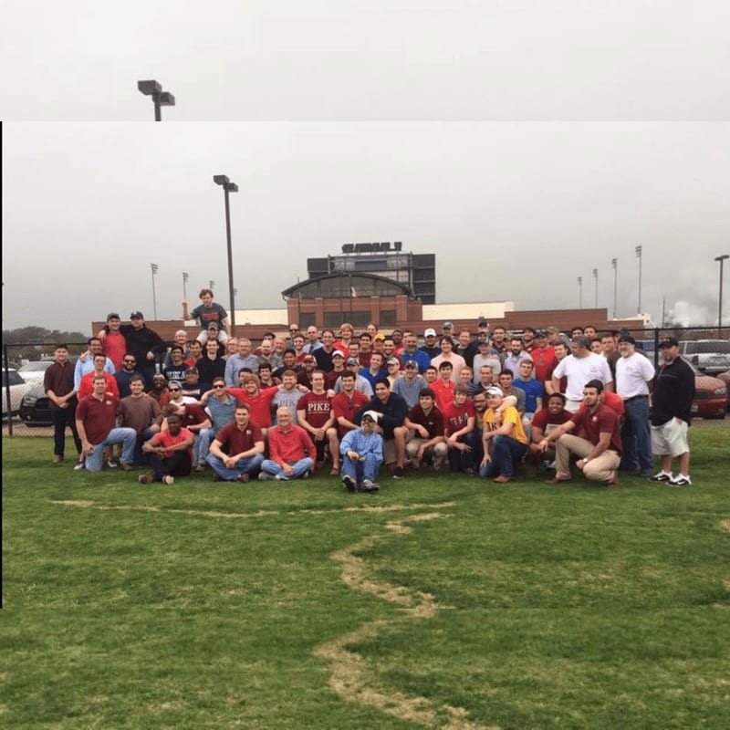 A large group of men in maroon shirts pose for a photo on a grassy field in front of a stadium under a cloudy sky.