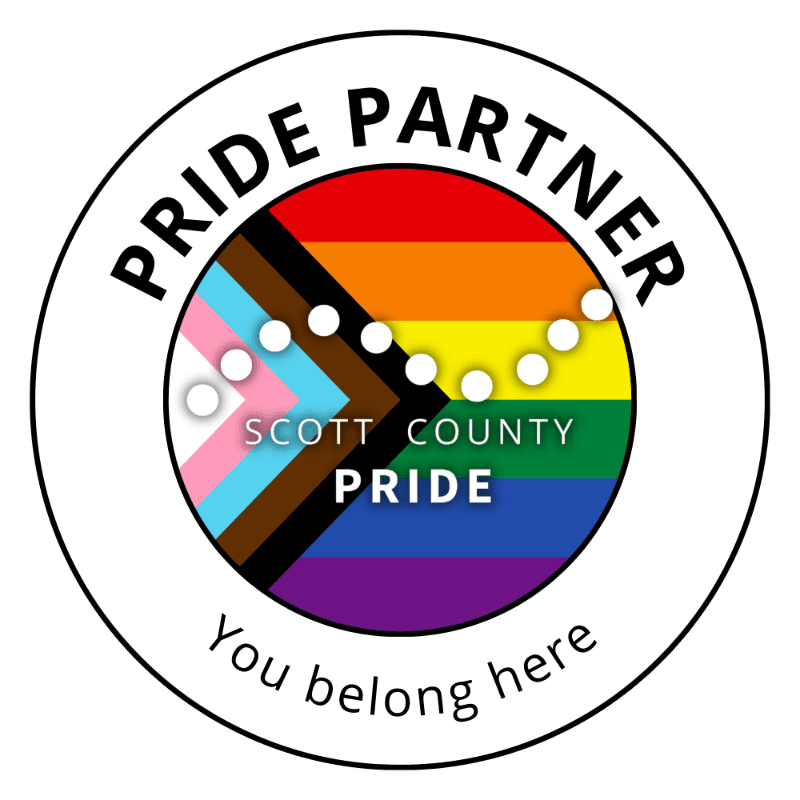 A circular logo with "PRIDE PARTNER" and "You belong here" text encircling a rainbow flag with the transgender pride flag overlayed, and "SCOTT COUNTY PRIDE" in the center.