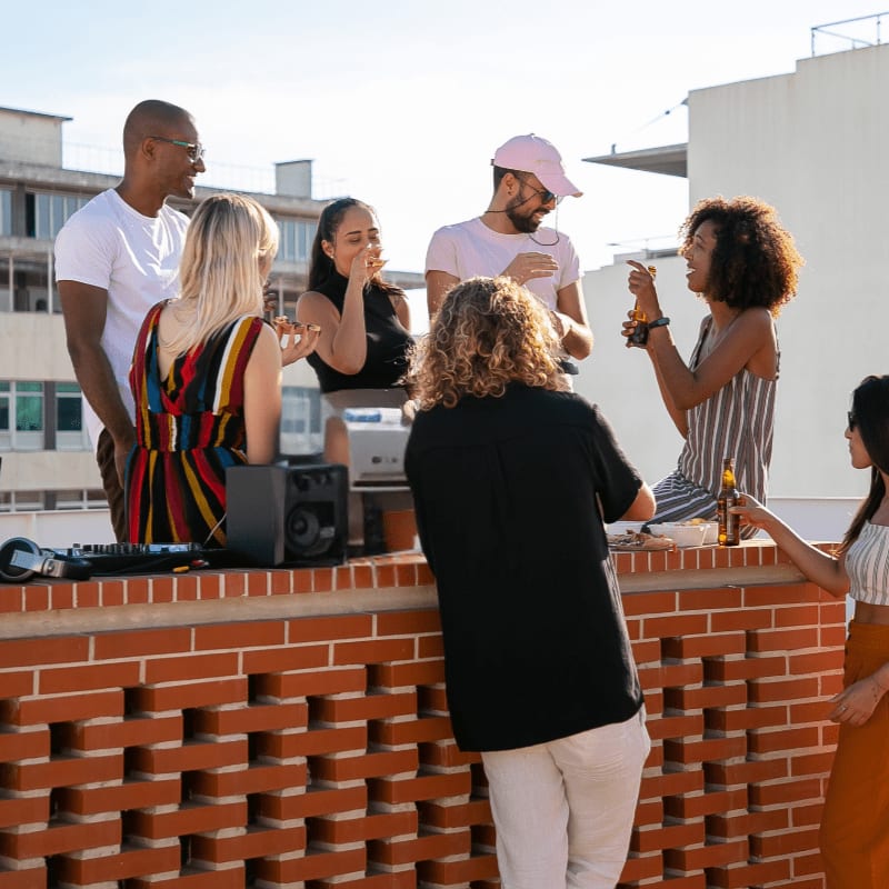 A group of friends enjoys a rooftop gathering with a DJ setup in the foreground and city buildings in the background.
