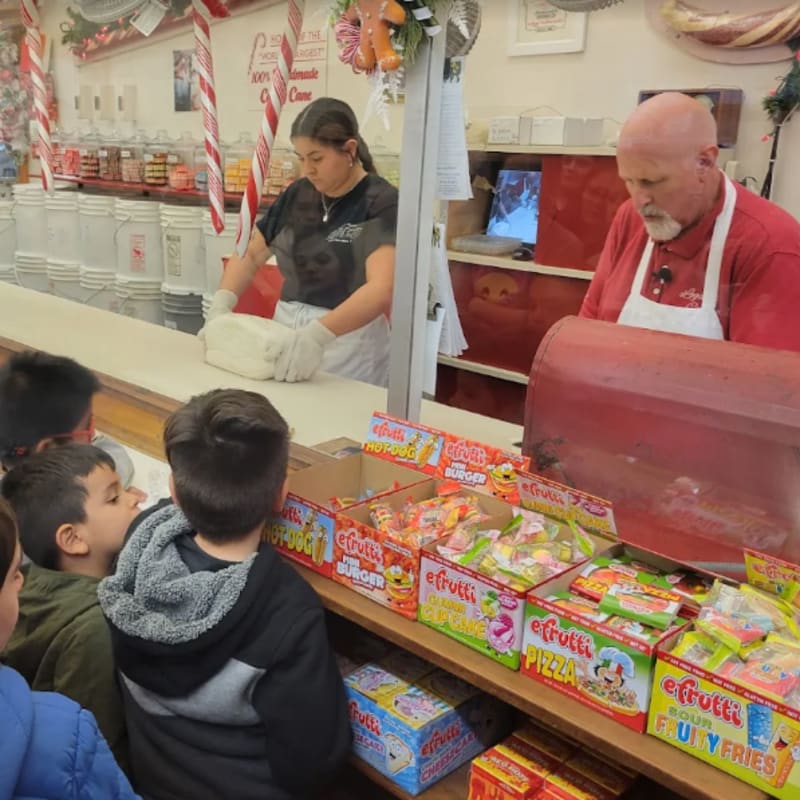 Children watch as a candy maker works taffy behind a counter filled with colorful candy boxes, with shelves of jars and candy canes visible in the background.