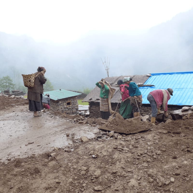 Several people are working on a muddy path in front of rustic houses, with misty mountains in the background.