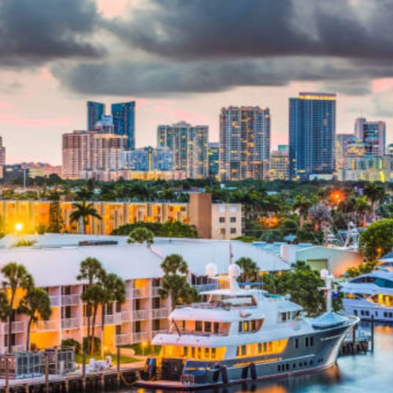 A large yacht is docked in a canal in the foreground, with a city skyline and palm trees in the background under a cloudy sky.