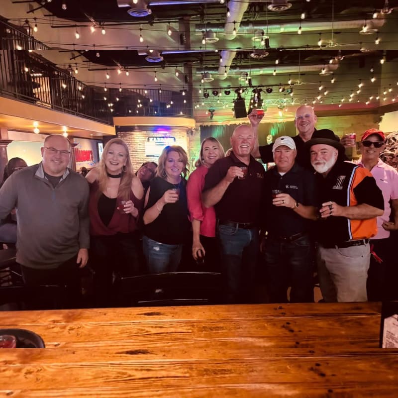 A group of smiling people stand behind a wooden bar in a dimly lit restaurant with string lights and a balcony in the background.