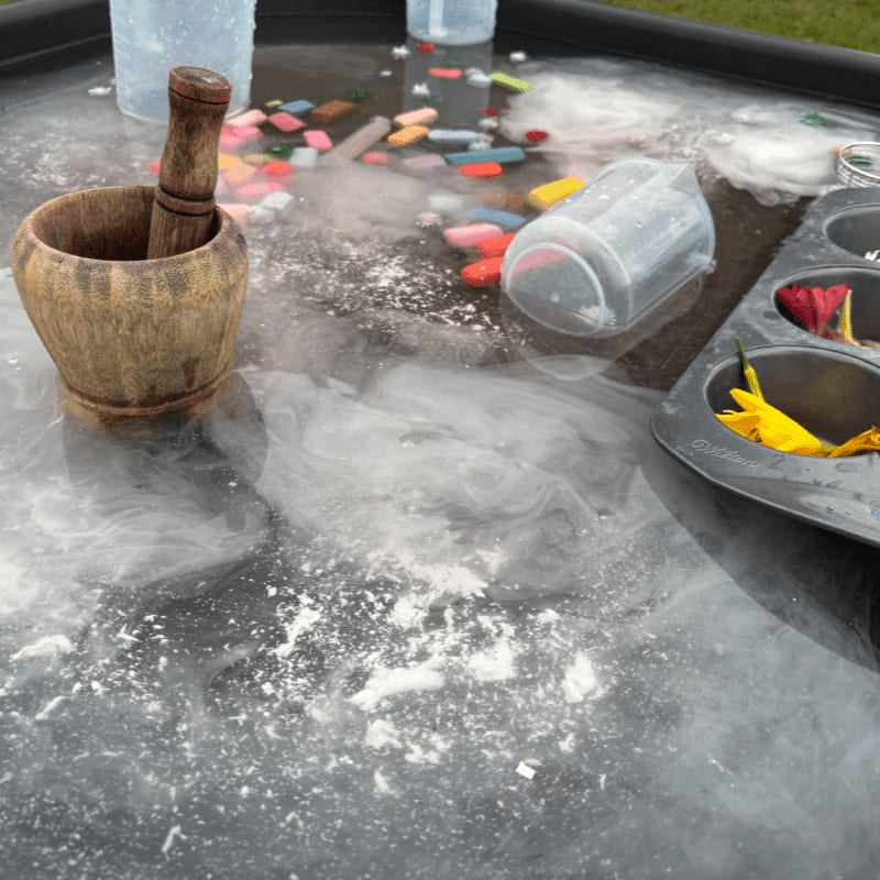A wooden mortar and pestle sit on a tray filled with colorful chalk pieces, water, and swirling mist, with a muffin tin holding flowers in the background.
