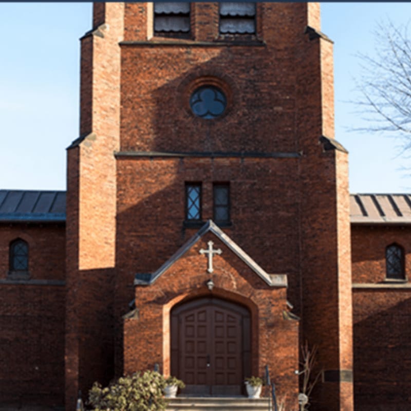 A brick church with a prominent tower and arched doorway stands against a clear blue sky, with bare tree branches visible to the right.