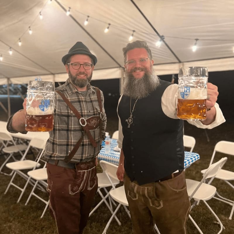 Two men in lederhosen and hats raise large beer steins in a tent decorated with string lights and checkered tablecloths.