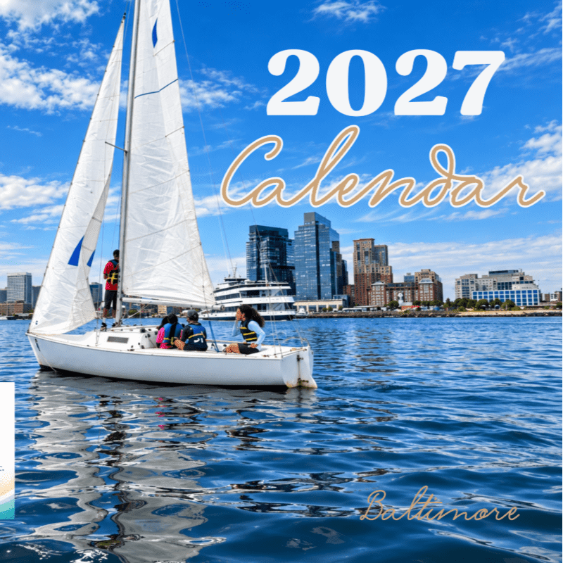 A sailboat with people aboard glides across blue water in the foreground, with a cityscape and a bright blue sky with clouds in the background.