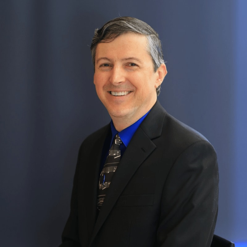 A smiling man in a suit is pictured in the foreground, with a plain blue background behind him.