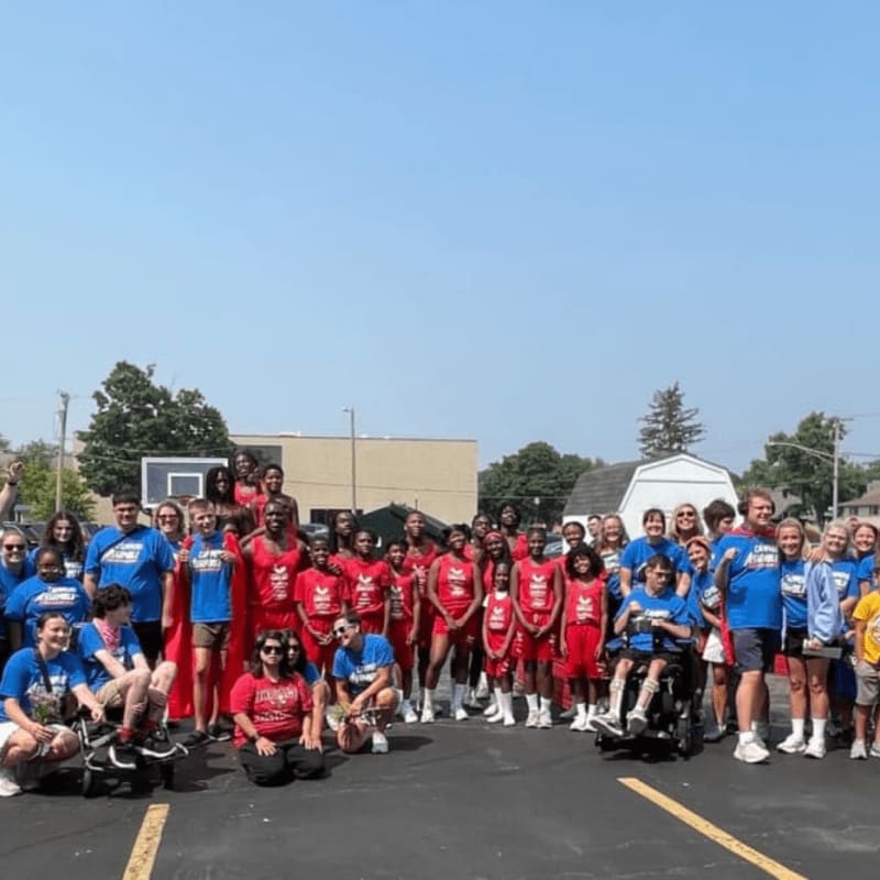 A large group of people, some in red athletic wear and others in blue t-shirts, pose for a photo in a parking lot with a building and trees in the background.