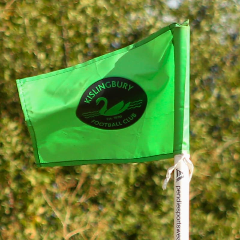 A bright green corner flag with the Kislingbury Football Club logo stands in the foreground against a blurred background of green foliage.