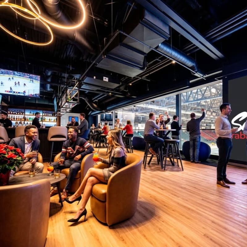 In the foreground, people relax in plush armchairs at a bar with drinks, while in the background, a crowd cheers at a sporting event visible through large windows.