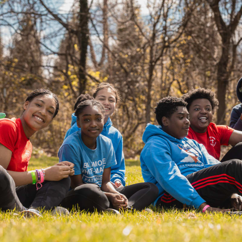 A group of smiling young people in casual clothing sit on a grassy field with trees in the background.