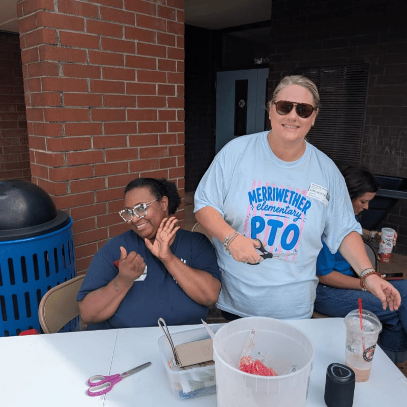 Two women smile at the camera from behind a table with various items, with a brick building and a blue trash can in the background.