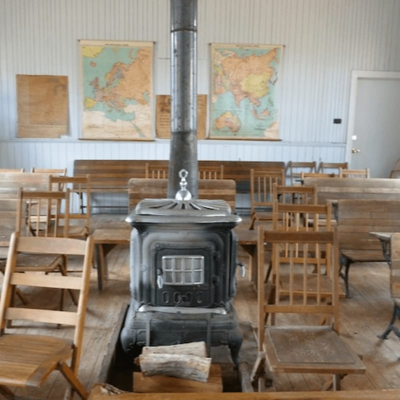 A rustic wood-burning stove sits in the foreground of a classroom with wooden desks and chairs, while vintage maps hang on the wall in the background.