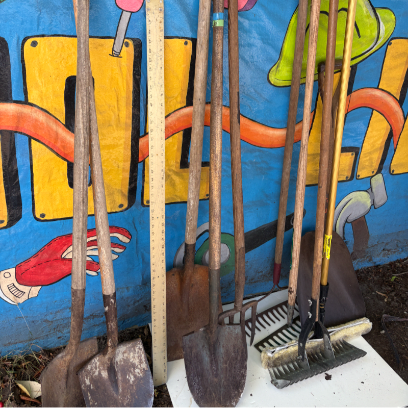 Several shovels and gardening tools stand in front of a colorful, cartoonish backdrop.
