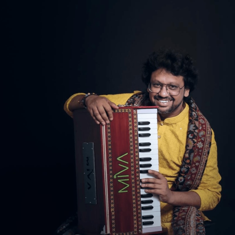 A smiling man in a yellow kurta and patterned shawl poses with a harmonium against a dark background.