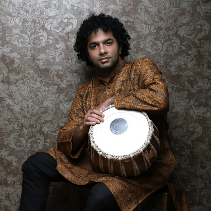 A man with curly hair in a traditional Indian outfit sits holding a tabla drum in front of a patterned wallpaper.
