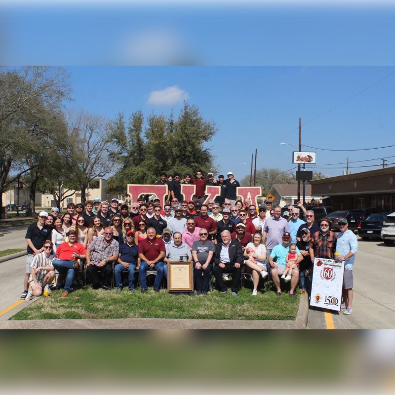 A large group of people pose for a photo in front of a giant "USA" sign, with a suburban street and buildings in the background.