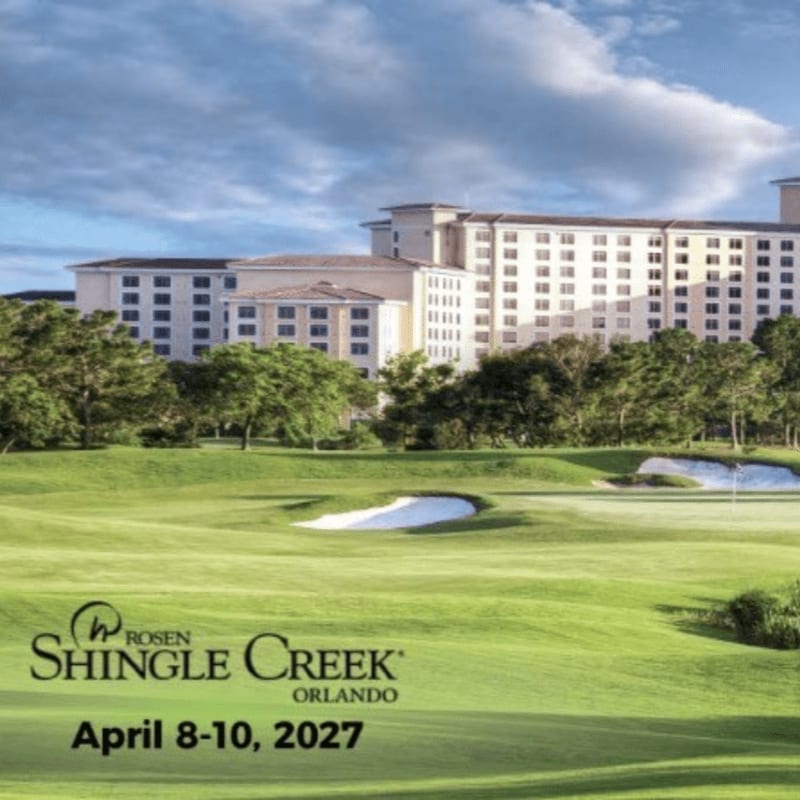 A lush green golf course with sand traps in the foreground leads to a large hotel complex under a cloudy sky in the background.